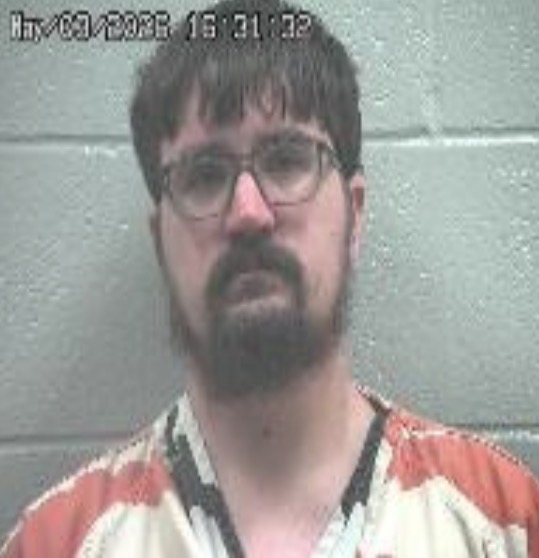 Mugshot of a man with glasses and a dark beard wearing an orange prison jumpsuit, standing against a gray cinder block wall; timestamp appears in the upper left.
