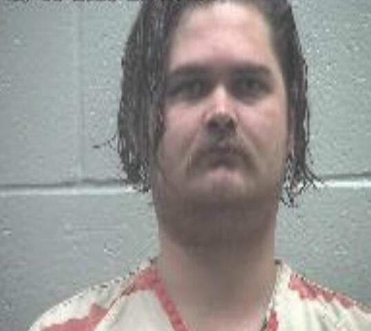 Mugshot of a man with medium-length dark hair and a mustache, wearing a red-and-white striped jail uniform, against a gray wall.