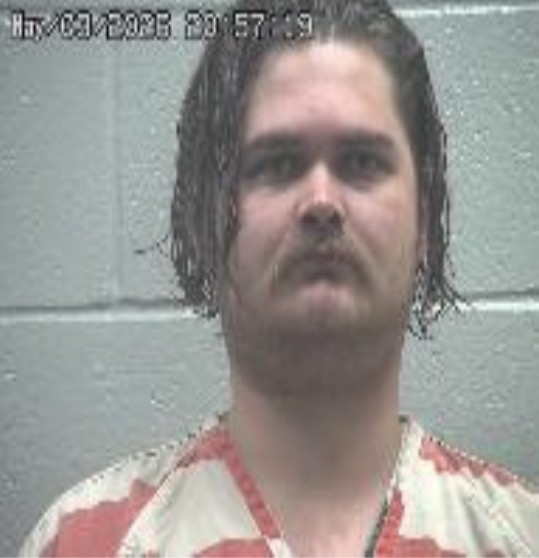Mugshot of a man with medium-length dark hair and a mustache, wearing a red-and-white striped jail uniform, against a gray wall.