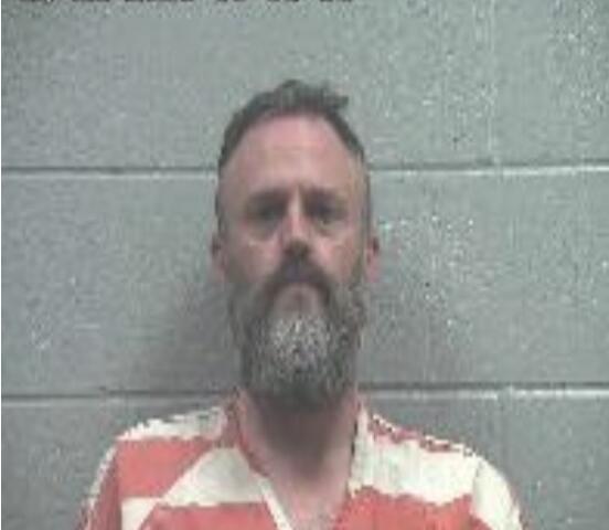 Mugshot of a middle-aged man with a gray beard wearing an orange striped jail uniform, standing in front of a gray cinder block wall; date/time stamp visible in the upper left corner.