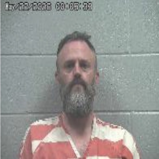 Mugshot of a middle-aged man with a gray beard wearing an orange striped jail uniform, standing in front of a gray cinder block wall; date/time stamp visible in the upper left corner.