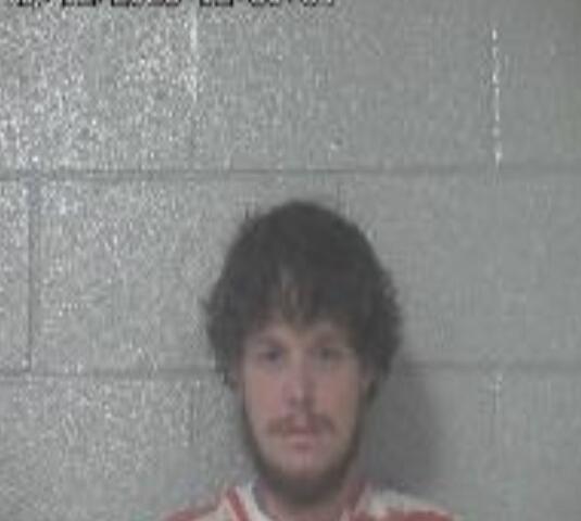 Mugshot of a man with dark hair and a beard in an orange jumpsuit against a gray cinder-block wall.
