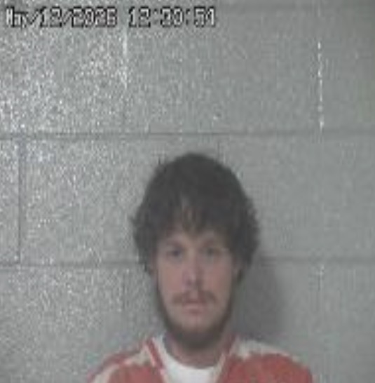 Mugshot of a man with dark hair and a beard in an orange jumpsuit against a gray cinder-block wall.