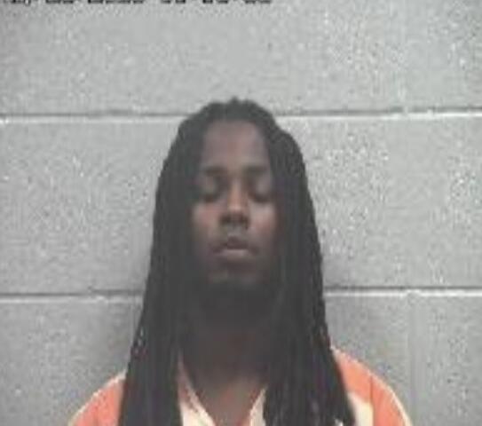 Mugshot of a man with long dreadlocks in an orange inmate jumpsuit, standing in front of a gray cinder block wall.