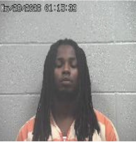 Mugshot of a man with long dreadlocks in an orange inmate jumpsuit, standing in front of a gray cinder block wall.
