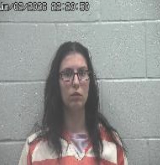 Mugshot of a woman with long dark hair and glasses, wearing an orange-and-white striped jail uniform, against a gray brick wall.