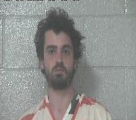 Mugshot of a bearded man in an orange-striped jail jumpsuit against a gray cinder block wall; timestamp visible in the top-left corner.