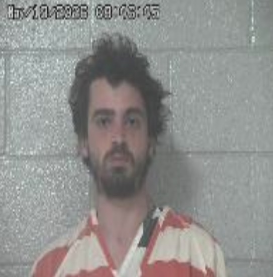 Mugshot of a bearded man in an orange-striped jail jumpsuit against a gray cinder block wall; timestamp visible in the top-left corner.