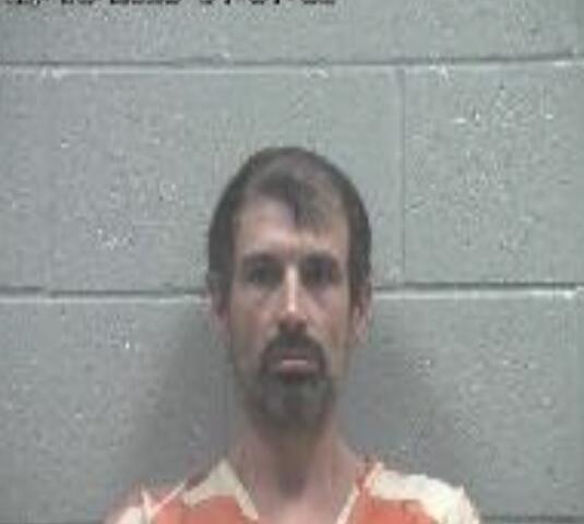 Mugshot of a man with short brown hair and goatee wearing an orange prison jumpsuit against a gray cinder block wall; date stamp visible at top.