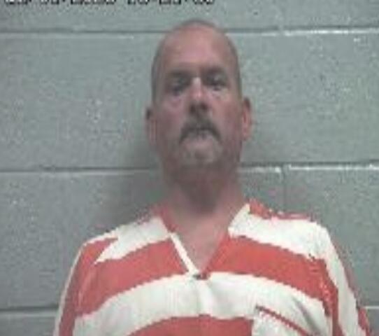 Mugshot of a middle-aged man with a mustache in an orange-and-white striped jail uniform, standing against a gray cinder block wall; timestamp in the upper-left corner.