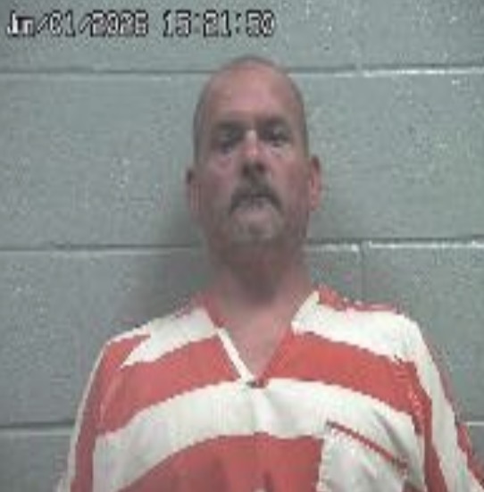 Mugshot of a middle-aged man with a mustache in an orange-and-white striped jail uniform, standing against a gray cinder block wall; timestamp in the upper-left corner.
