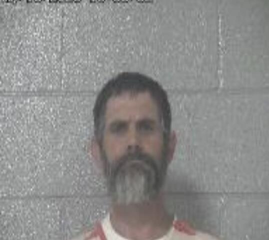 Mugshot of a man with dark hair and a gray beard standing against a gray cinder block wall.