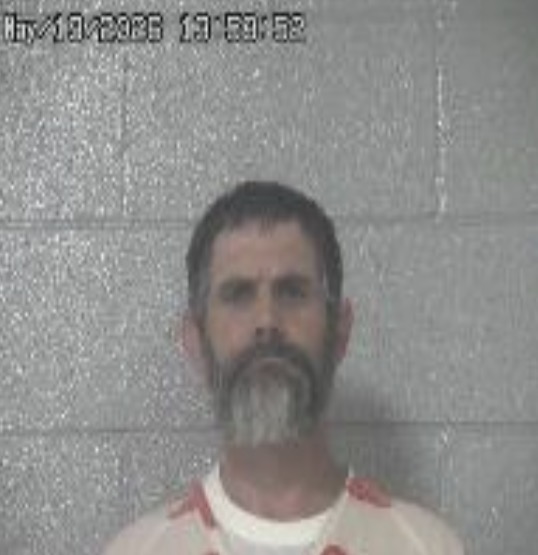 Mugshot of a man with dark hair and a gray beard standing against a gray cinder block wall.