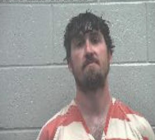 Mugshot of a man with dark hair and a goatee in an orange striped inmate jumpsuit, against a gray cinder block wall.