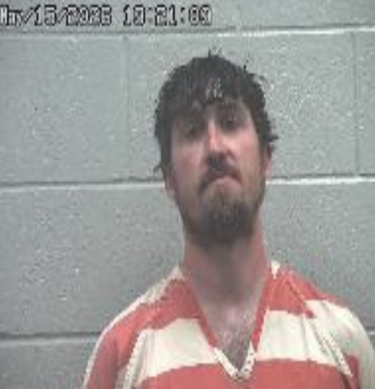 Mugshot of a man with dark hair and a goatee in an orange striped inmate jumpsuit, against a gray cinder block wall.