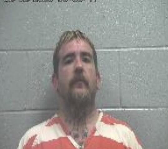 Mugshot of a man with blond hair and a goatee, wearing an orange-and-white striped prison uniform, standing against a gray cinder block wall; timestamp in the upper left reads 10:53:07.