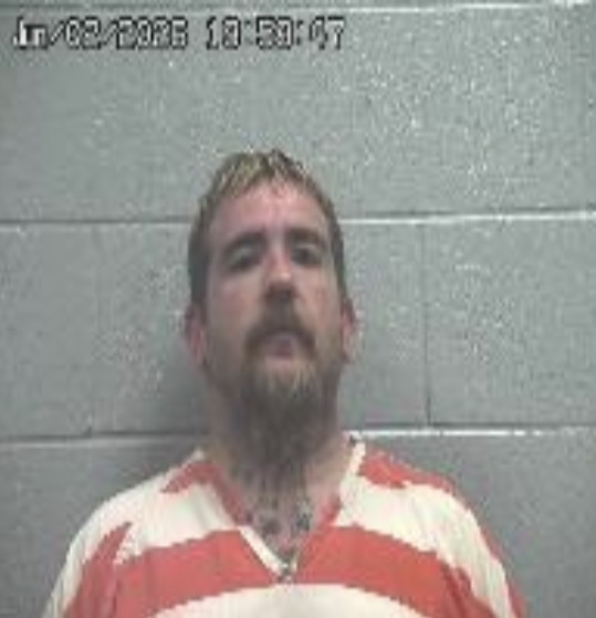 Mugshot of a man with blond hair and a goatee, wearing an orange-and-white striped prison uniform, standing against a gray cinder block wall; timestamp in the upper left reads 10:53:07.