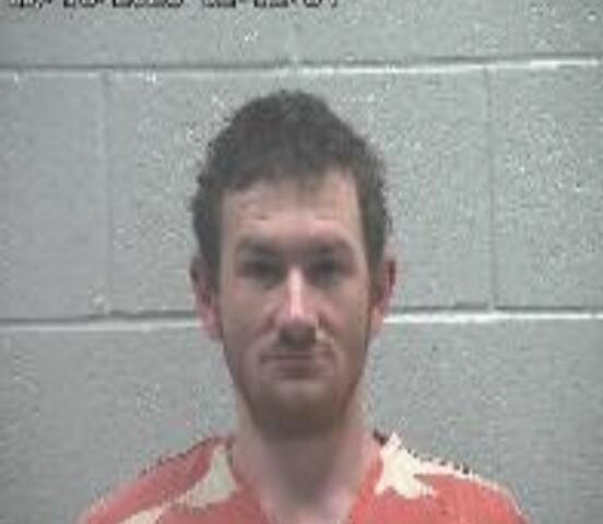 Mugshot-style photo of a man with short dark hair wearing orange clothing with white patterns, standing against a gray cinder block wall; timestamp appears in the top-left corner.