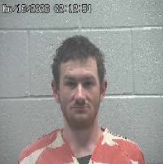 Mugshot-style photo of a man with short dark hair wearing orange clothing with white patterns, standing against a gray cinder block wall; timestamp appears in the top-left corner.