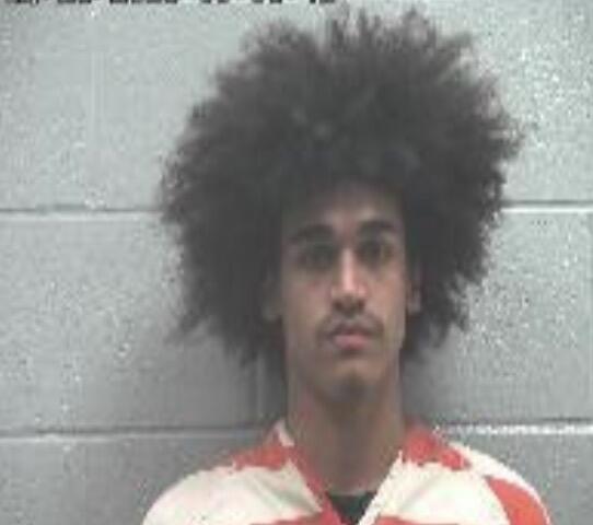 Mugshot of a young man with a large Afro against a gray cinder block wall, wearing an orange-and-white striped jail jumpsuit.