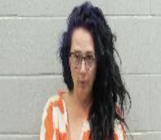 Portrait of a woman with long dark hair and glasses wearing an orange-striped jumpsuit, standing against a gray wall.