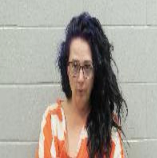 Portrait of a woman with long dark hair and glasses wearing an orange-striped jumpsuit, standing against a gray wall.