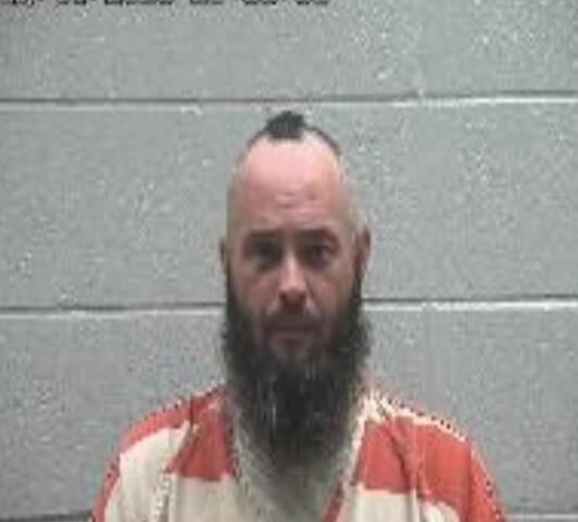 Mugshot of a man with a mohawk and full beard in an orange-and-white jail uniform, standing against a gray cinder block wall.