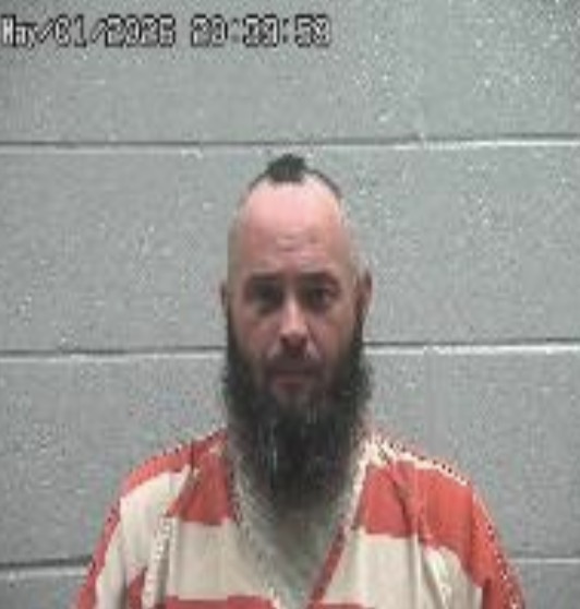 Mugshot of a man with a mohawk and full beard in an orange-and-white jail uniform, standing against a gray cinder block wall.