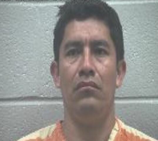 Mugshot of a man in an orange striped prison jumpsuit facing the camera against a gray cinder block wall.