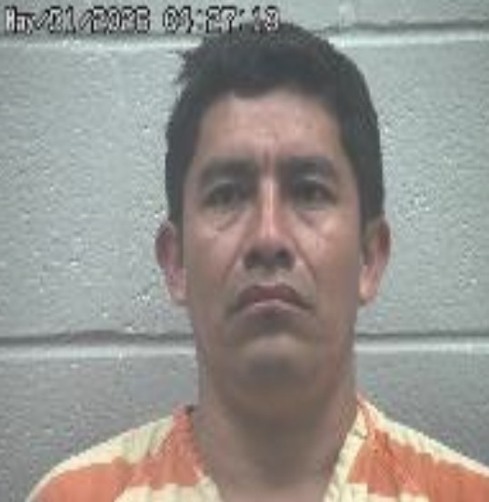 Mugshot of a man in an orange striped prison jumpsuit facing the camera against a gray cinder block wall.