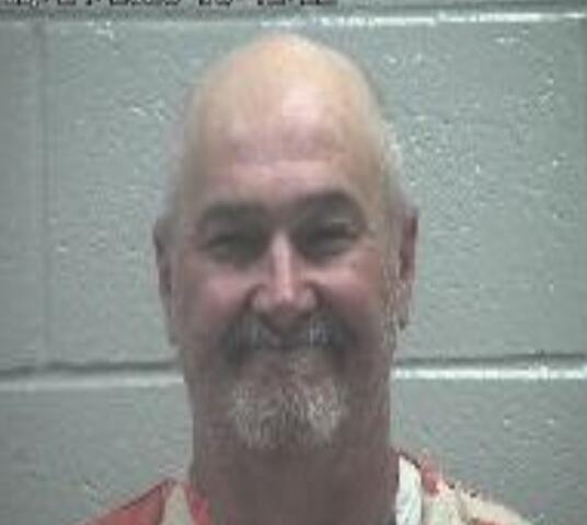 Smiling, bald man with a gray goatee in an orange prison jumpsuit, standing in front of a gray cinder‑block wall; appears to be a mugshot.