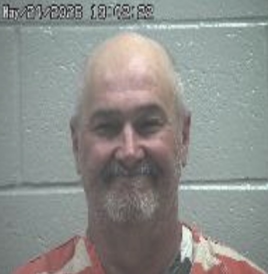Smiling, bald man with a gray goatee in an orange prison jumpsuit, standing in front of a gray cinder‑block wall; appears to be a mugshot.