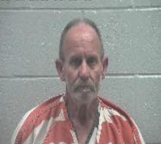 Mugshot of an older man with a gray goatee and receding hairline, wearing a red-and-white striped prison jumpsuit, against a gray cinder block wall (timestamp overlay at top).