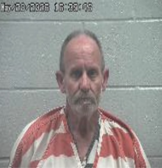 Mugshot of an older man with a gray goatee and receding hairline, wearing a red-and-white striped prison jumpsuit, against a gray cinder block wall (timestamp overlay at top).