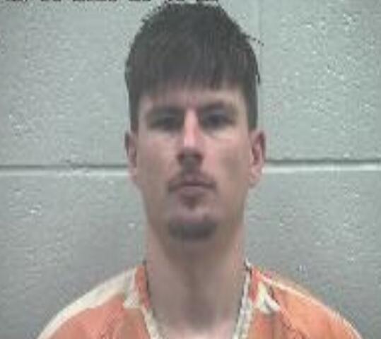 Mugshot of a man with short dark hair wearing an orange jail jumpsuit against a gray wall with a timestamp at the top left.