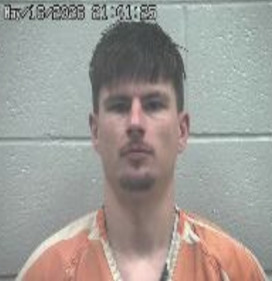 Mugshot of a man with short dark hair wearing an orange jail jumpsuit against a gray wall with a timestamp at the top left.
