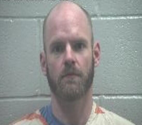 Mugshot photo of a bald man with a short beard, wearing a light striped jumpsuit, standing against a gray cinder block wall.