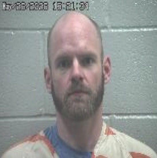 Mugshot photo of a bald man with a short beard, wearing a light striped jumpsuit, standing against a gray cinder block wall.