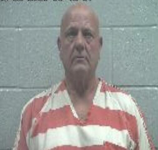 Mugshot of an older man with a shaved head in orange-and-white striped inmate clothing, posed against a gray wall.
