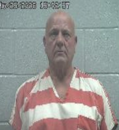Mugshot of an older man with a shaved head in orange-and-white striped inmate clothing, posed against a gray wall.