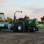Green tractor pulling a weight-sled on a dirt track at a rural tractor-pull event; a man sits at the controls behind the machine.