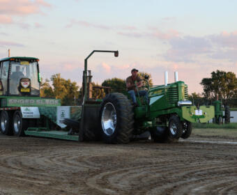 Green tractor pulling a weight-sled on a dirt track at a rural tractor-pull event; a man sits at the controls behind the machine.