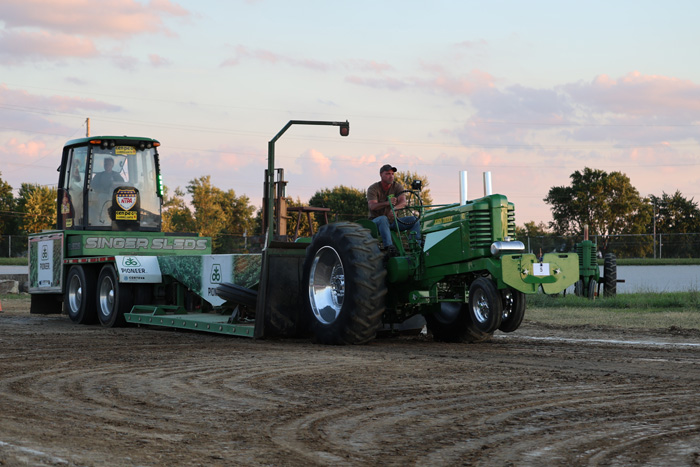 Green tractor pulling a weight-sled on a dirt track at a rural tractor-pull event; a man sits at the controls behind the machine.