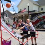 Two boys play a basketball shooting game on a street fair, one about to release a ball toward the hoop, with a crowd and a truck in the background.