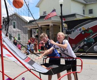 Two boys play a basketball shooting game on a street fair, one about to release a ball toward the hoop, with a crowd and a truck in the background.