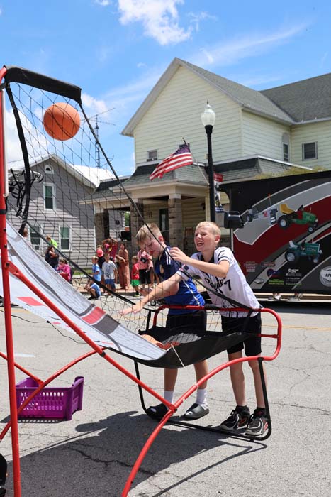 Two boys play a basketball shooting game on a street fair, one about to release a ball toward the hoop, with a crowd and a truck in the background.