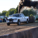 White pickup towing a trailer with a heavy vehicle at a dirt track, black smoke rising from the exhaust as spectators watch.