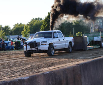White pickup towing a trailer with a heavy vehicle at a dirt track, black smoke rising from the exhaust as spectators watch.