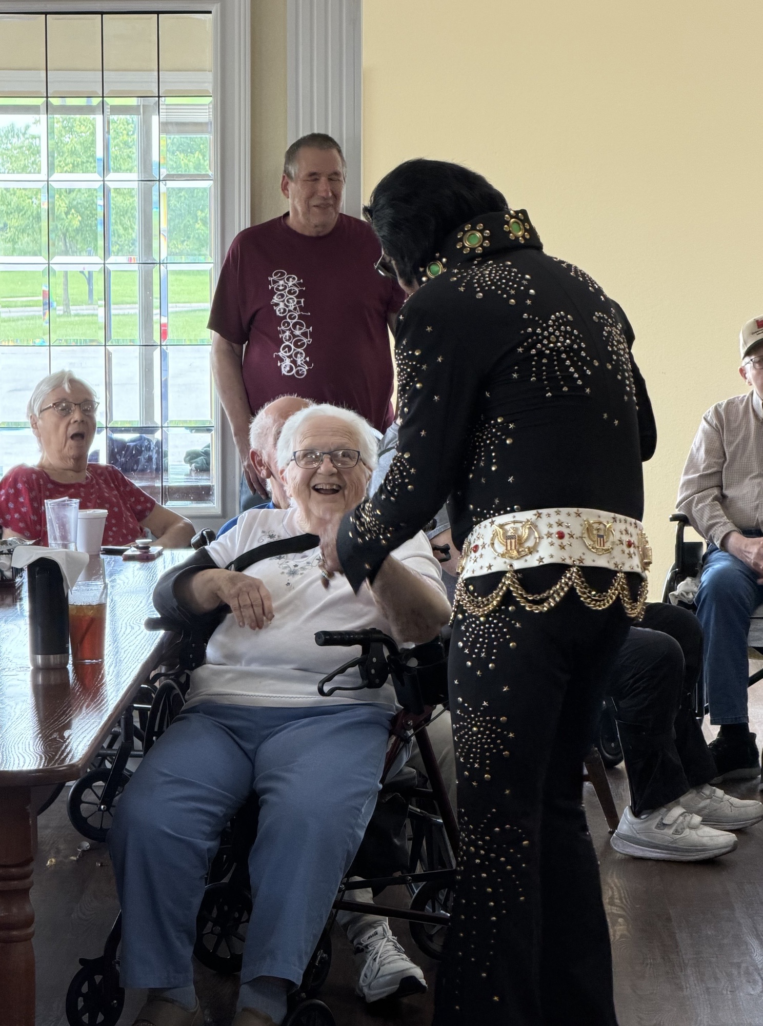 Elderly man in a wheelchair at a dining table, smiling as a performer in a black studded jumpsuit leans in nearby in a bright room with large windows behind them.