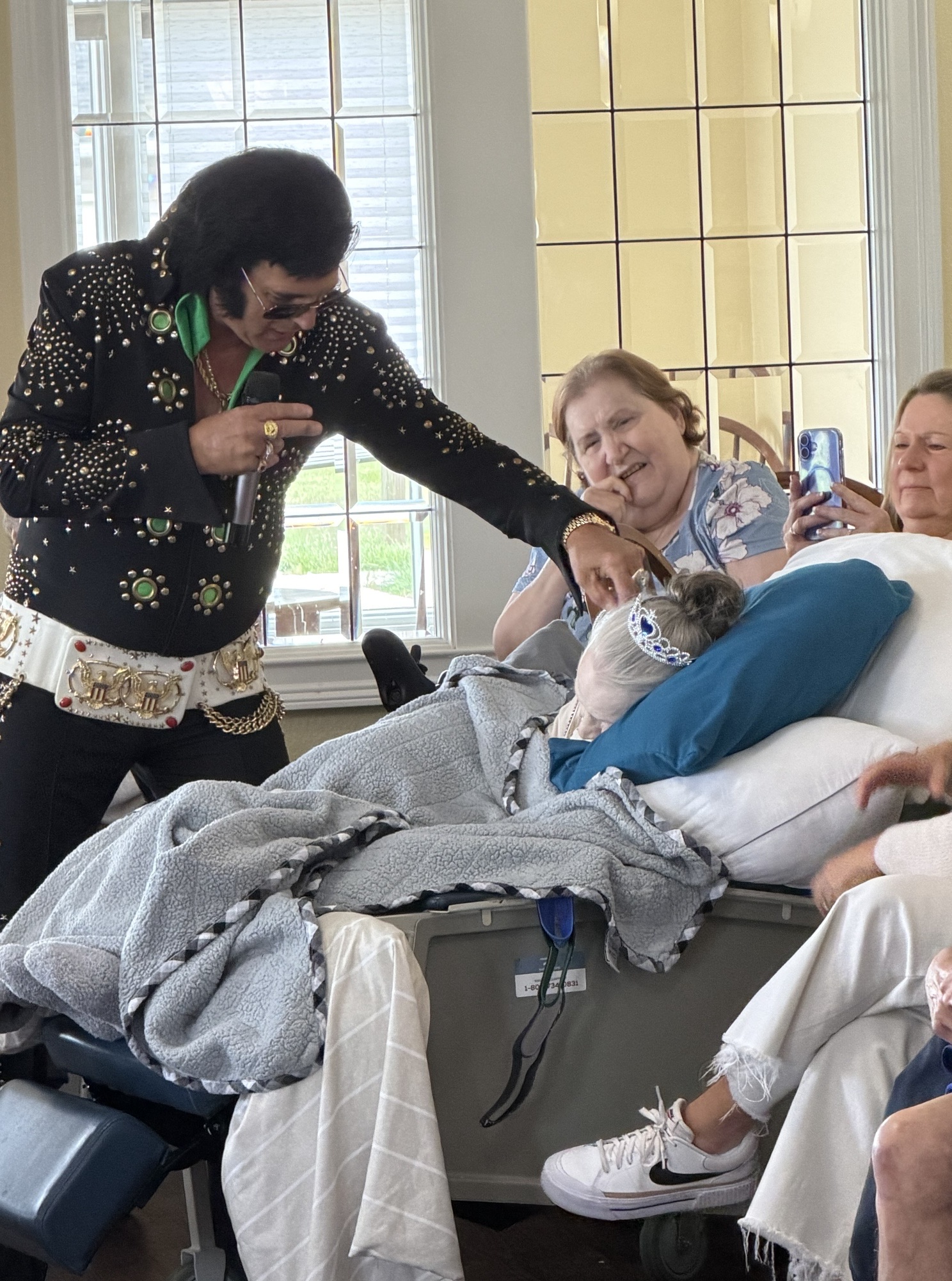 Elvis-style performer in a studded jacket sings to elderly patients in a hospital room as a woman nearby records on her phone.